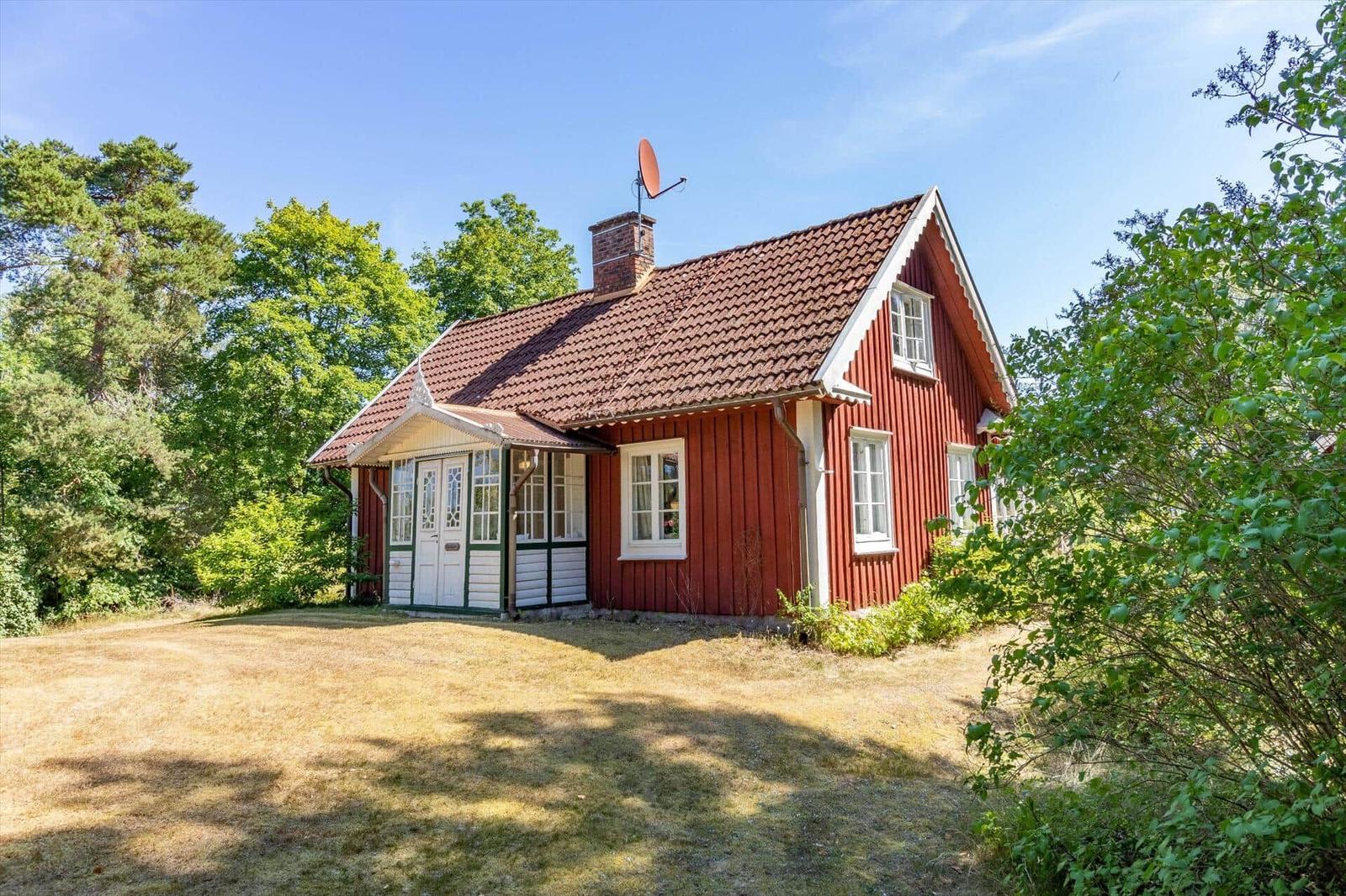 Rotes Holzhaus mit Dach und Antenne, umgeben von Grünflächen und Bäumen.