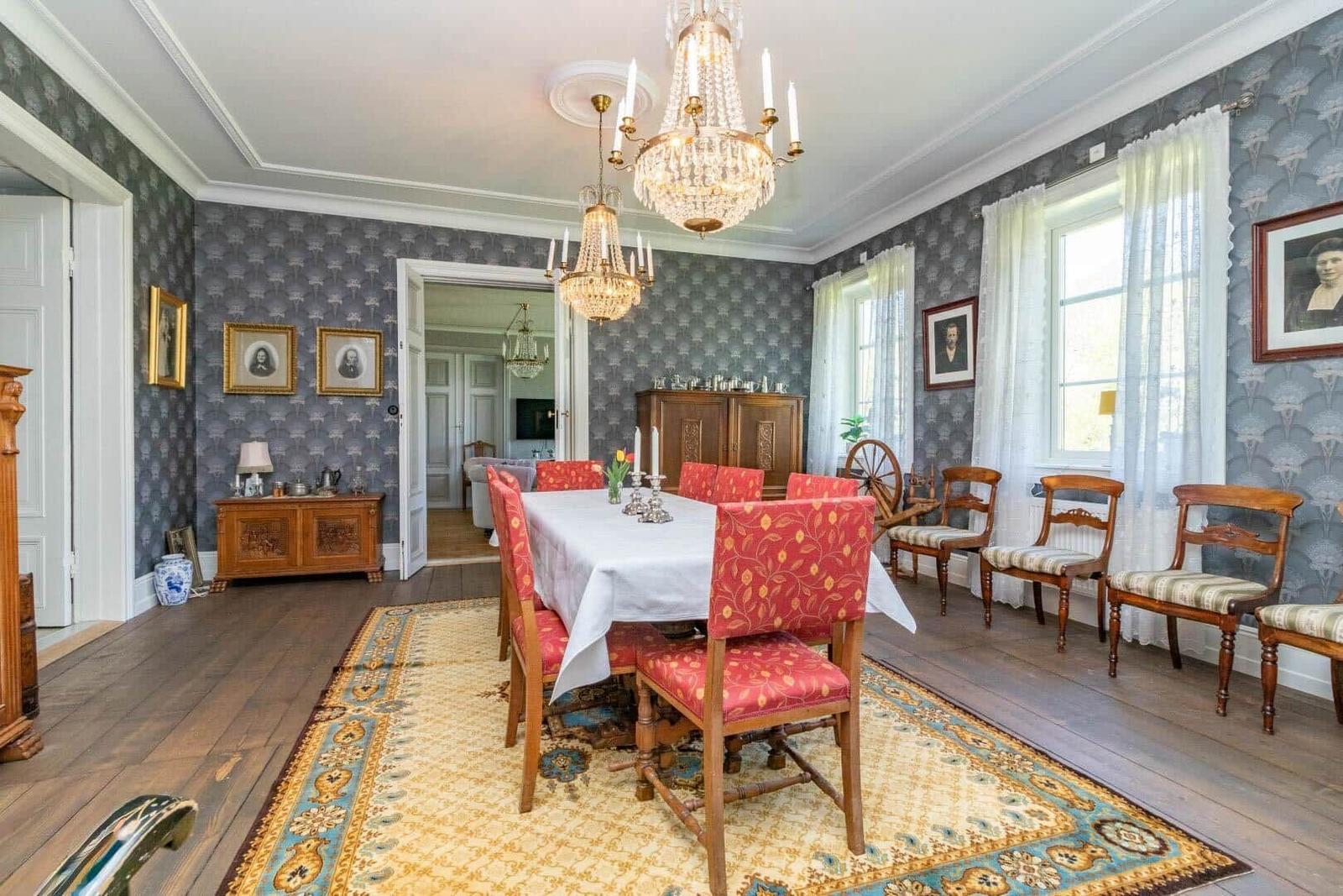 Esszimmer mit Holzboden, gemustertem Teppich und Kronleuchtern. An den Wänden hängen Porträts.