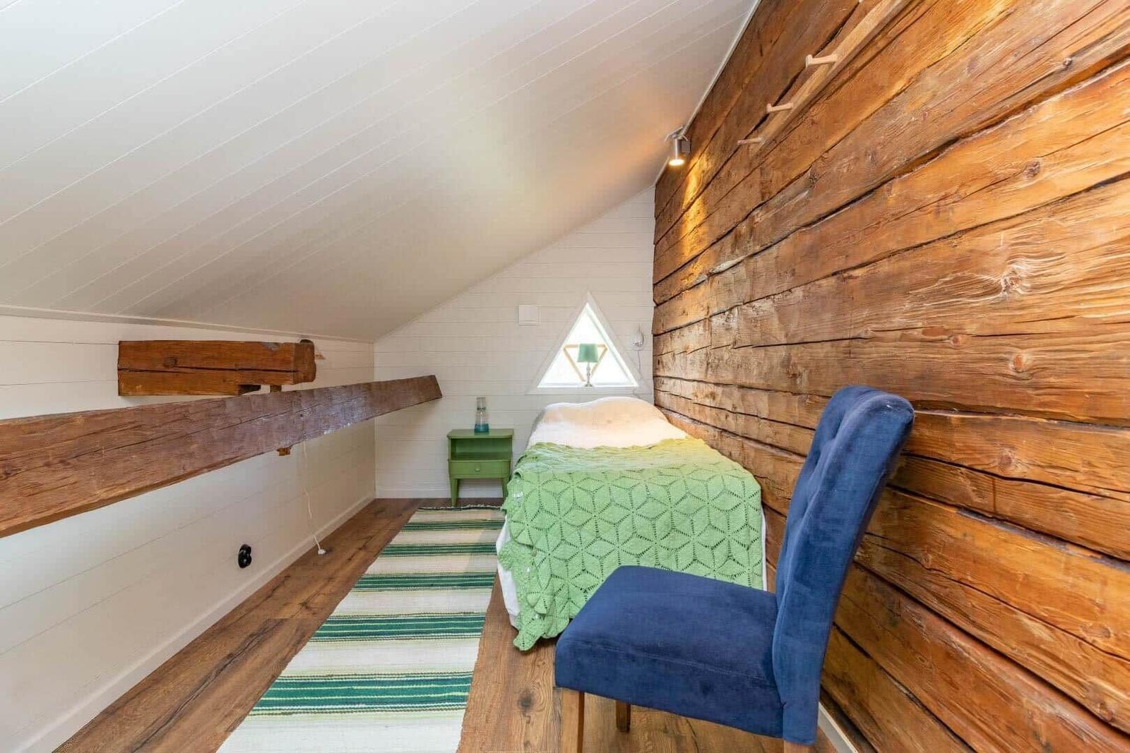 Schlafzimmer mit Holzwand, Bett, Stuhl und Dreiecksfenster unter Dachschräge.