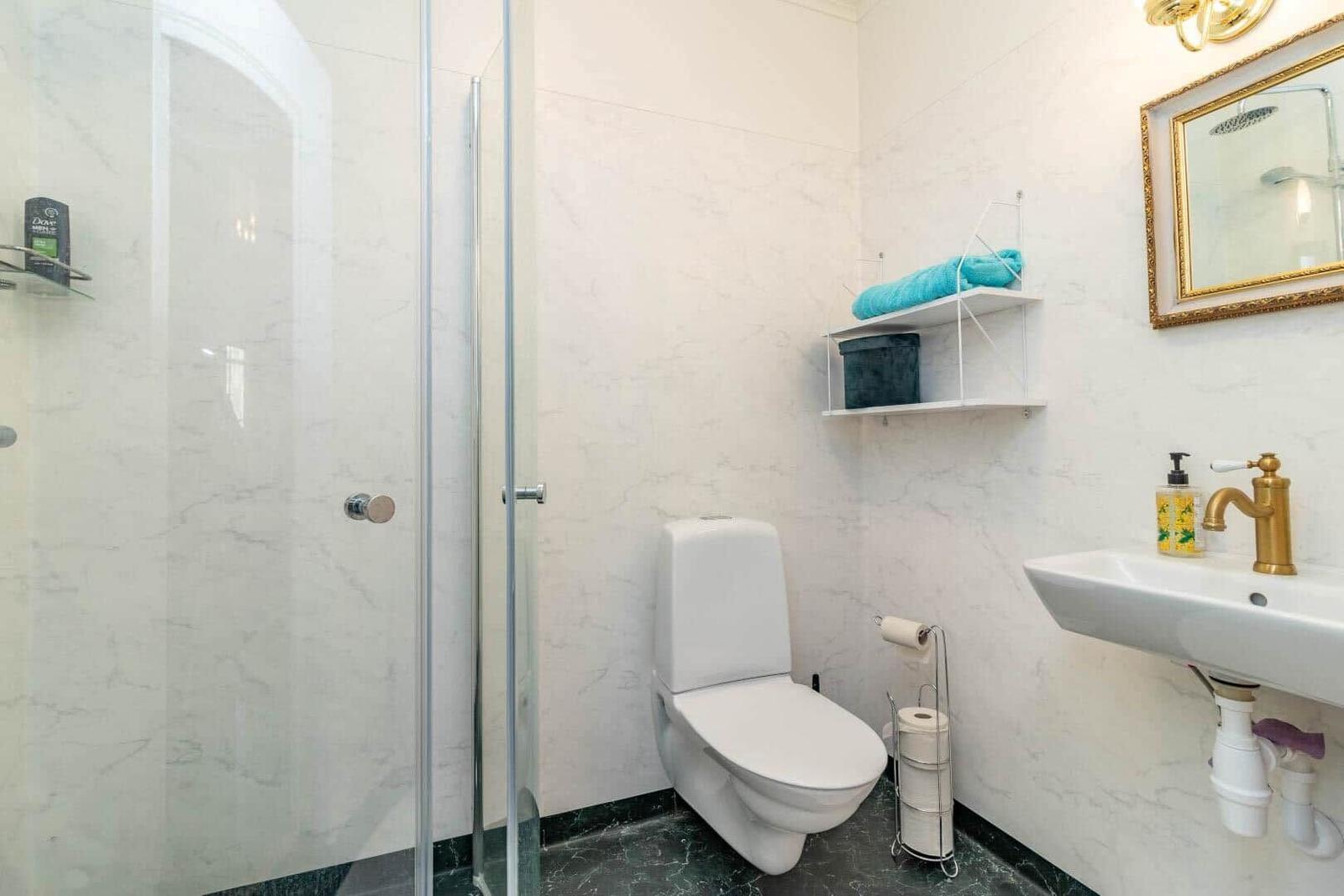 Badezimmer mit Dusche, Toilette und Waschbecken. Weiße Wände und goldener Wasserhahn.