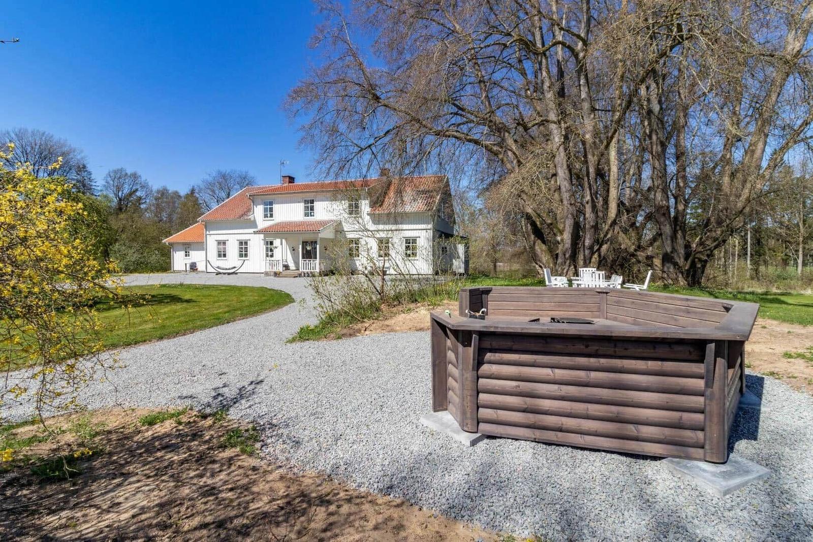 Ein weißes Haus mit rotem Dach, umgeben von Gras und Bäumen. Ein Holzbad steht auf Kies.