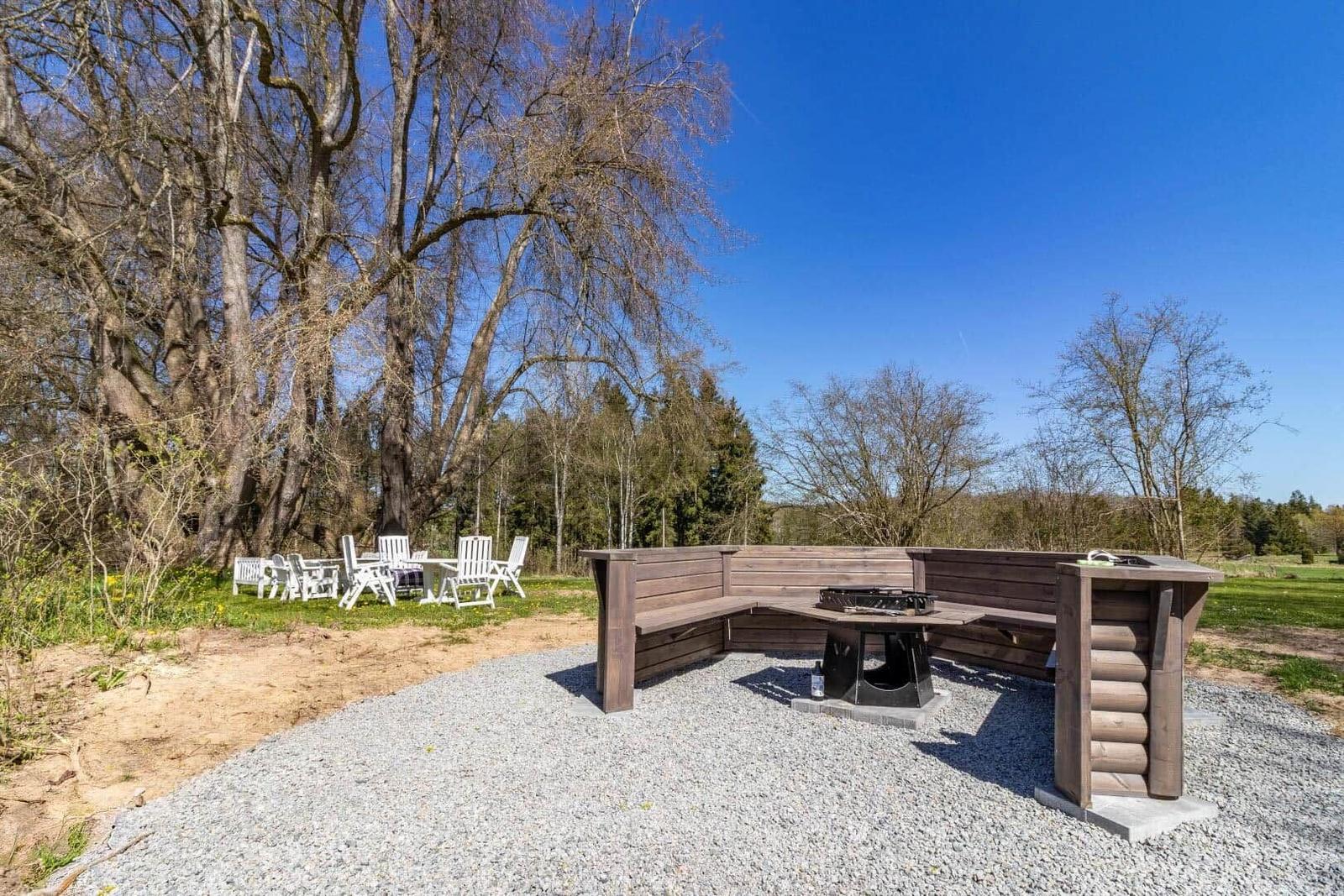 Holzbank mit Grill auf Kiesfläche, weiße Gartenmöbel unter Bäumen.
