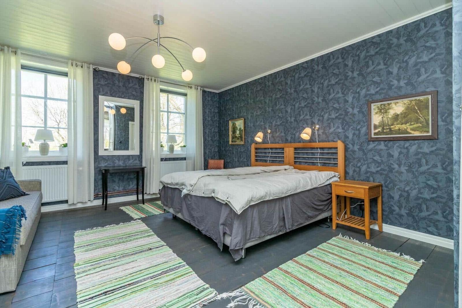 Schlafzimmer mit Bett, zwei Fenstern, Wandbildern und einem modernen Deckenlicht.