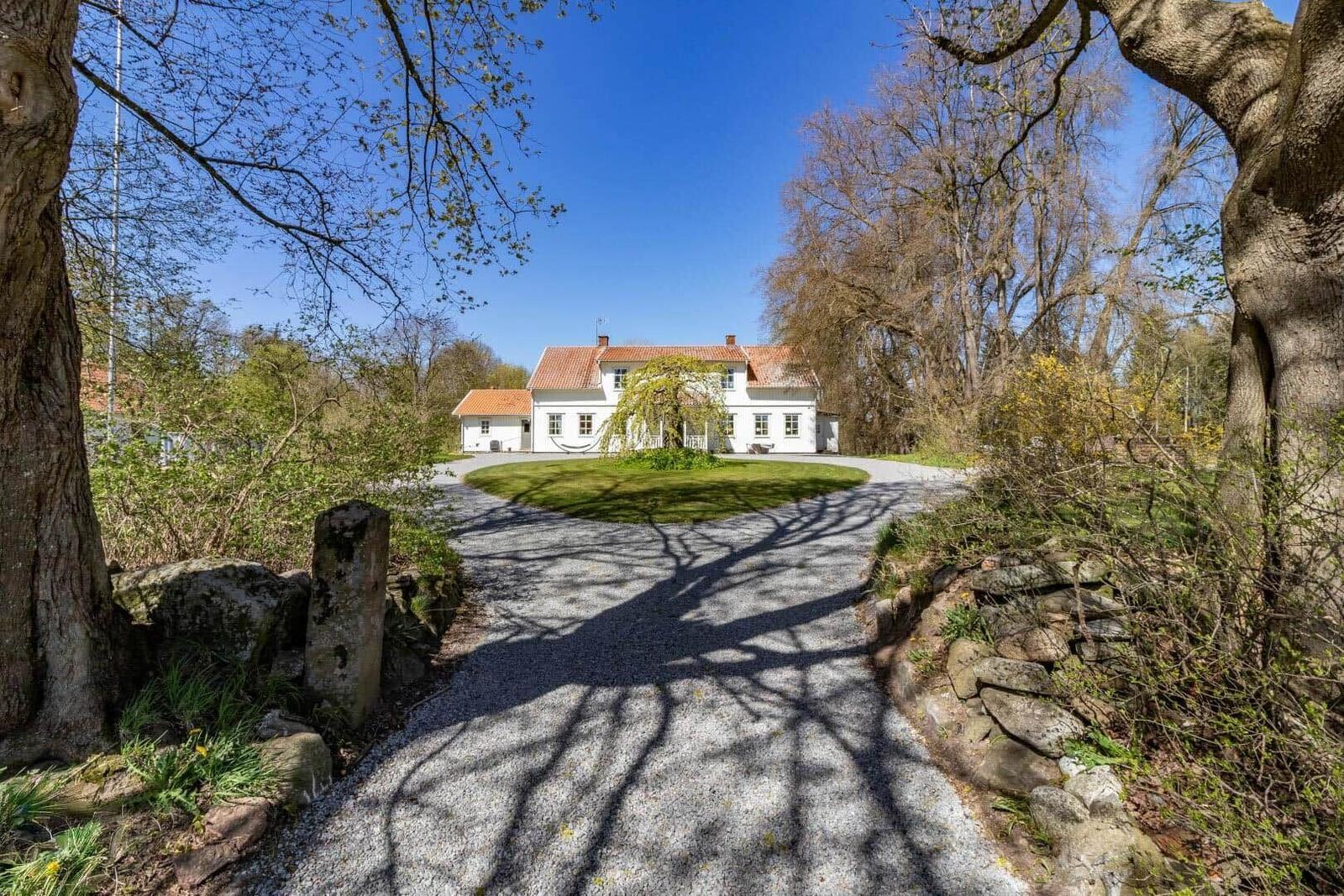 Ein weißes Haus mit rotem Dach steht hinter einem Kiesweg, umgeben von Bäumen und Gras.