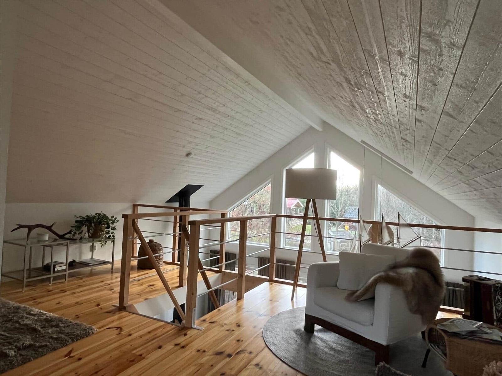 Ein Wohnbereich mit Holzboden, weißer Couch und großen Fenstern unter einem geneigten Dach.
