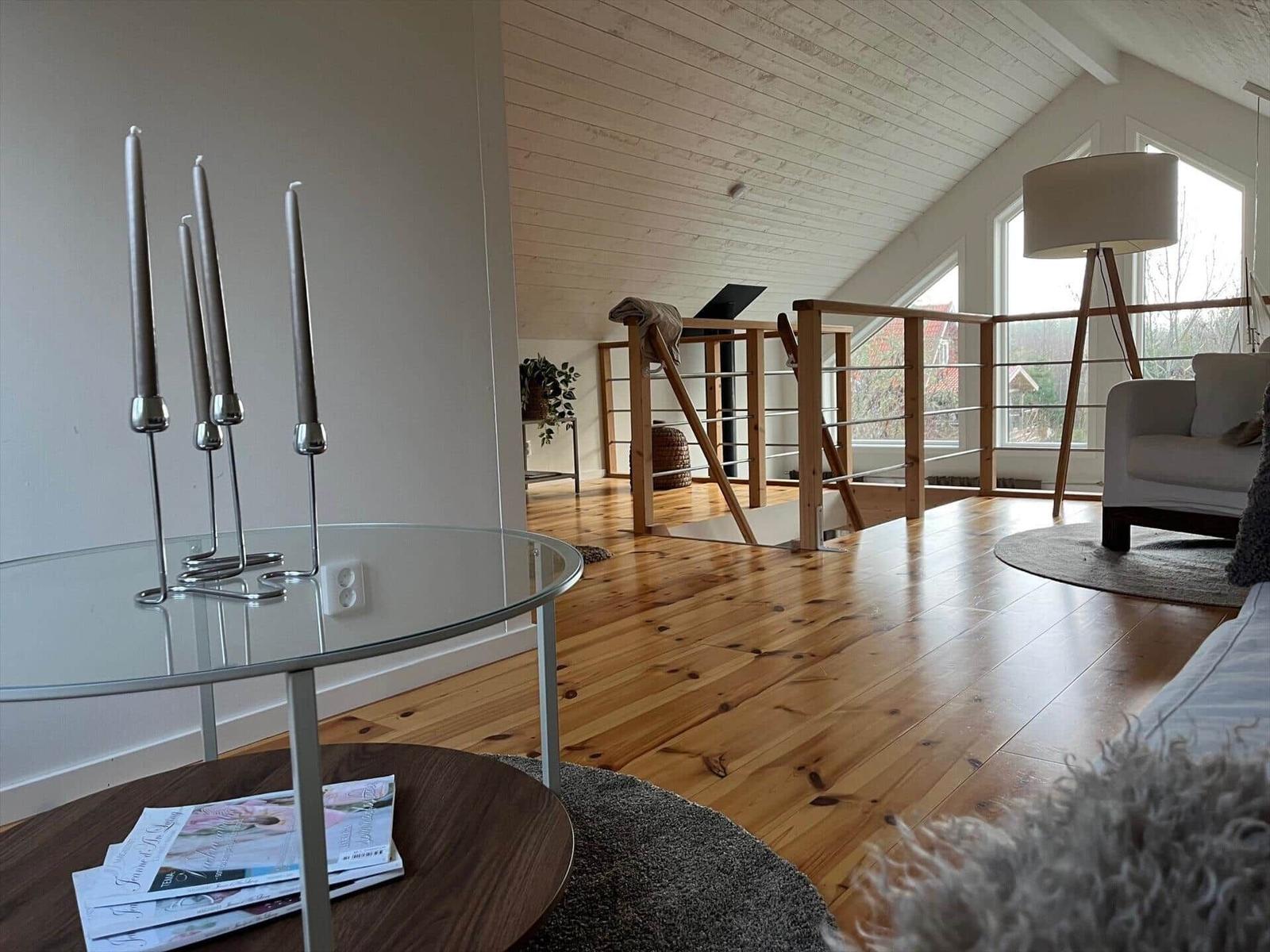 Wohnbereich mit Holzboden, Glas-Tisch und Sitzgelegenheit. Holzgeländer und große Fenster.