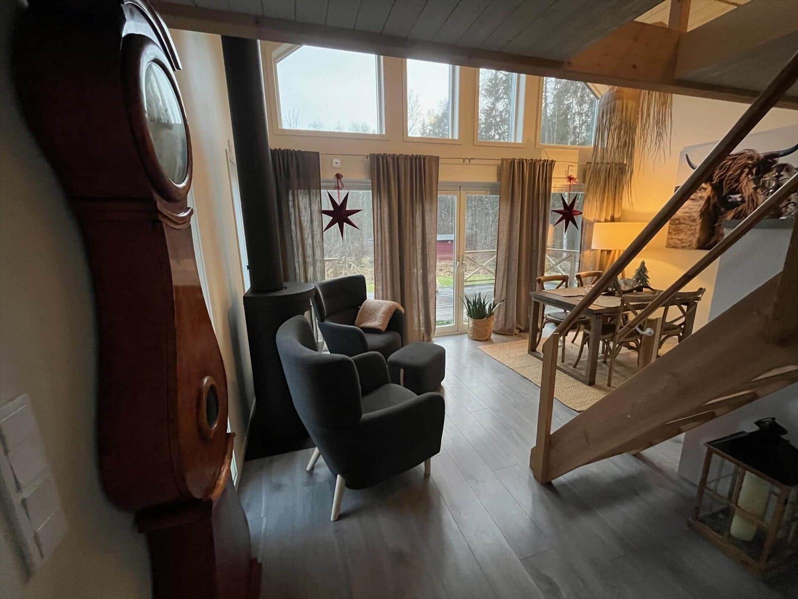 Wohnraum mit Sitzgelegenheiten, Essbereich und Holztreppe. Große Fenster und Holzdecke.