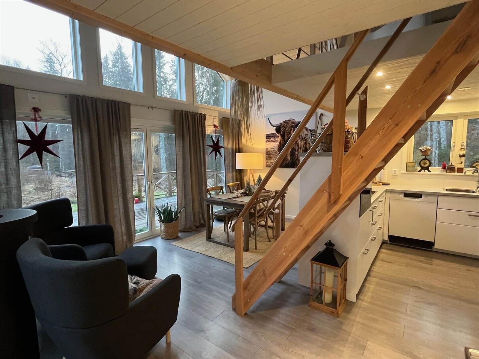 Wohn- und Essbereich mit Küche und Holztreppe. Große Fenster und Türen zu Terrasse.