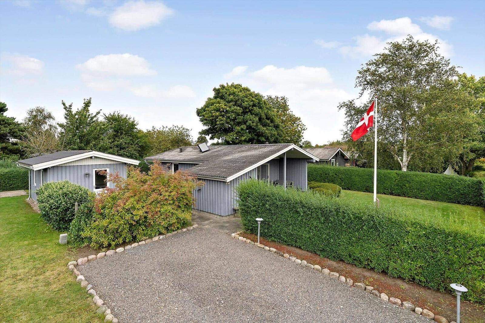 Ein graues Ferienhaus mit Garten, Hecke und dänischer Flagge. 