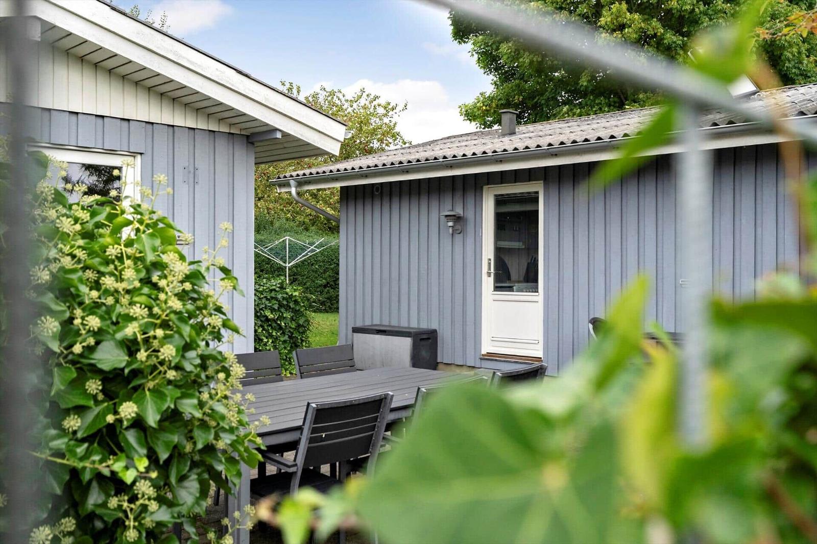 Haus mit Terrasse, Tisch und Stühlen. Umgeben von Grün und Pflanzen.