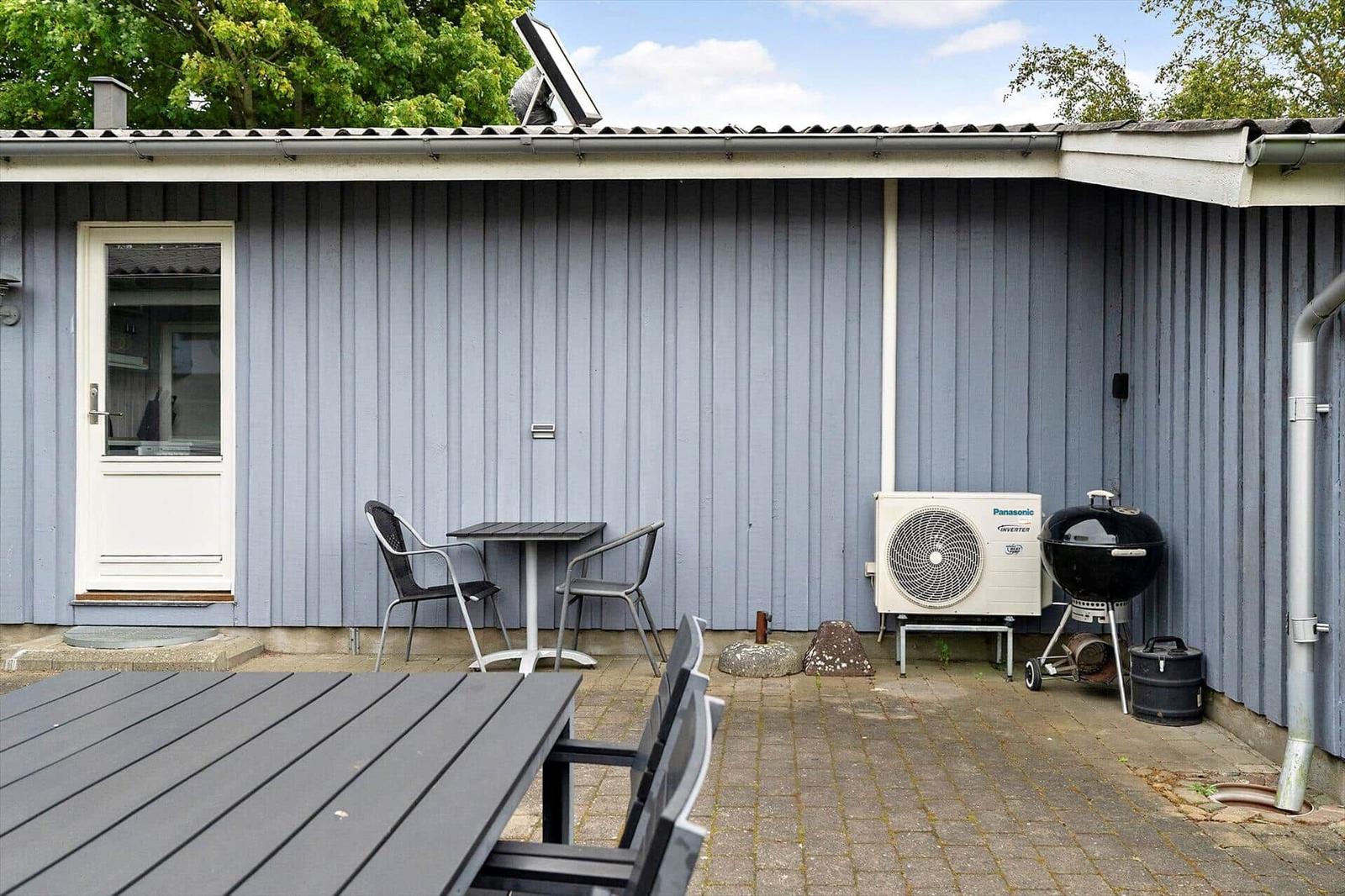 Terrasse mit Tisch, Stühlen, Grill und Klimaanlage an grauer Holzwand.