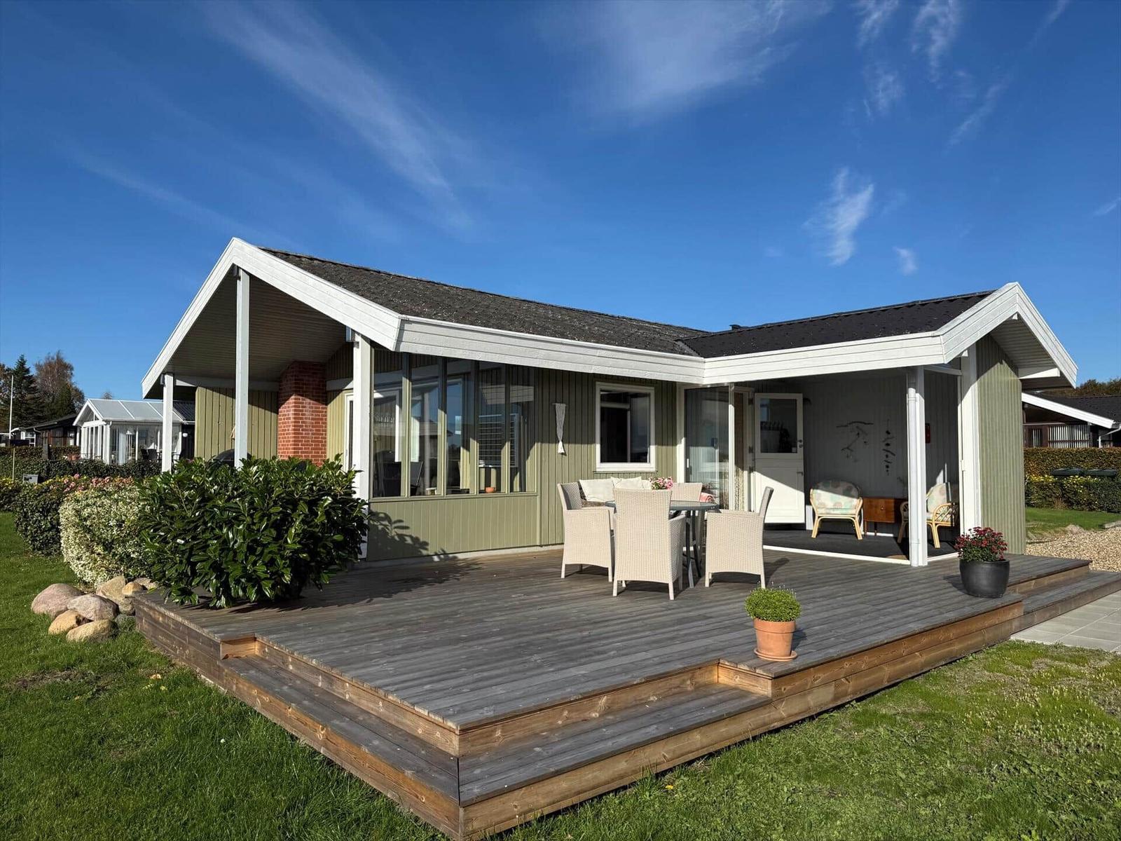 Haus mit großem Holzdeck, Sitzgelegenheiten und Garten. Helle Fenster und moderne Architektur.