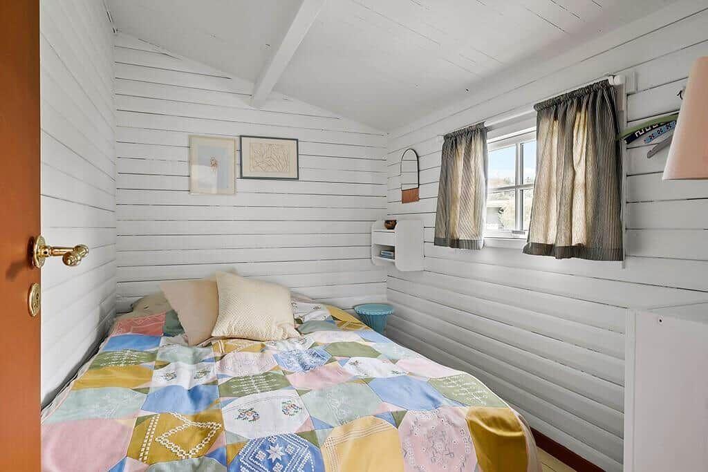Bedroom with white paneled walls, bed with colorful quilt, and window with curtains.