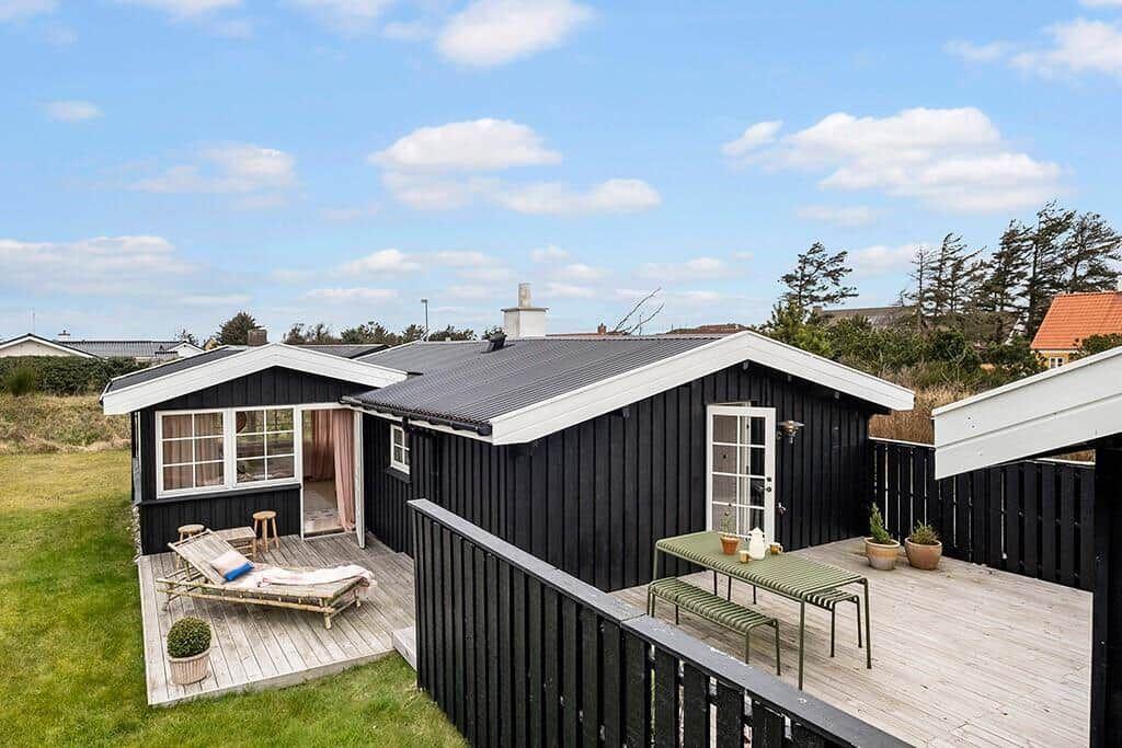 Black wooden house with terrace, lounge chair, and table. Green lawn and blue sky.