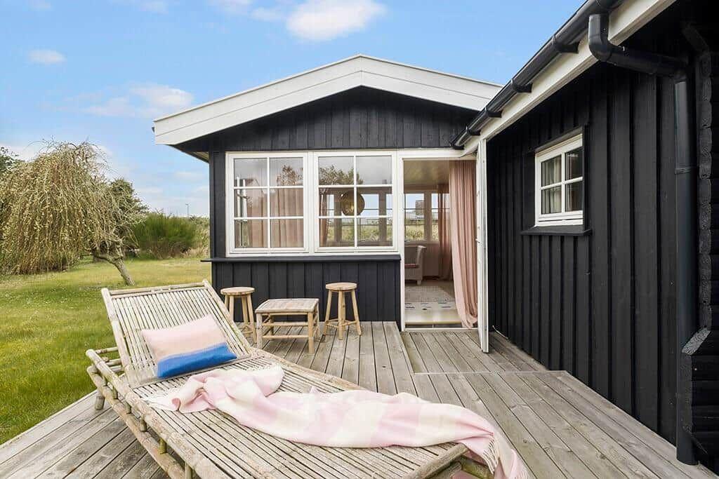 Wooden terrace with lounge chair, table and stools in front of black wooden house with windows.