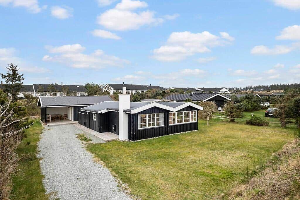 Black wooden house with white chimney and terrace. Lawn and gravel path.