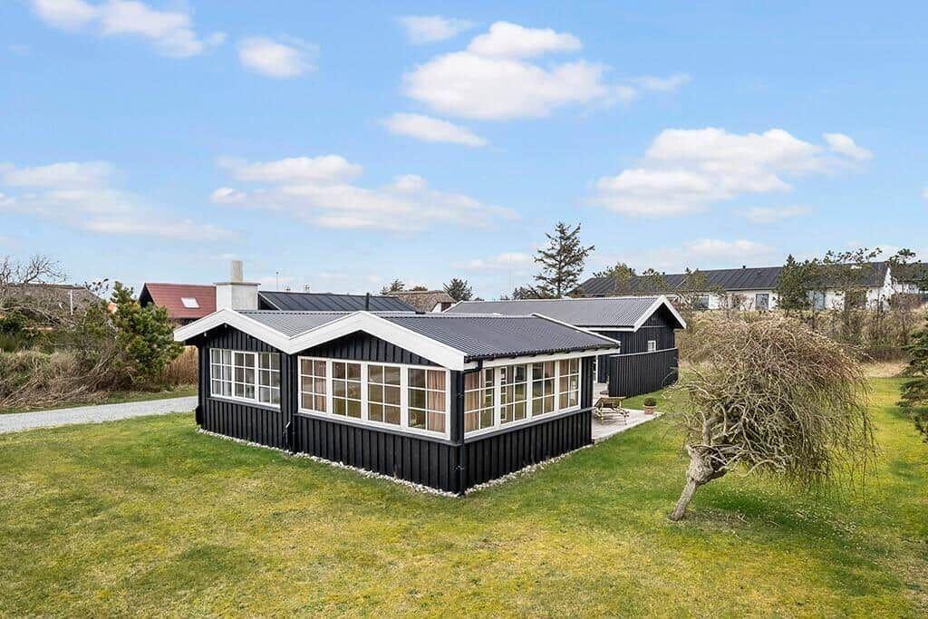 Black wooden house with large windows and garden. Background shows other houses.