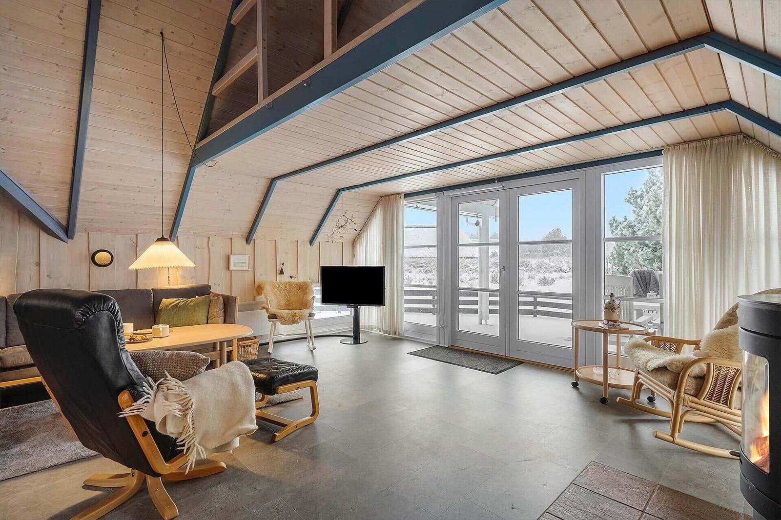 Wohnzimmer mit Holzwand, Fernseher, Sessel und Kamin. Große Fenster mit Blick auf Außenbereich.