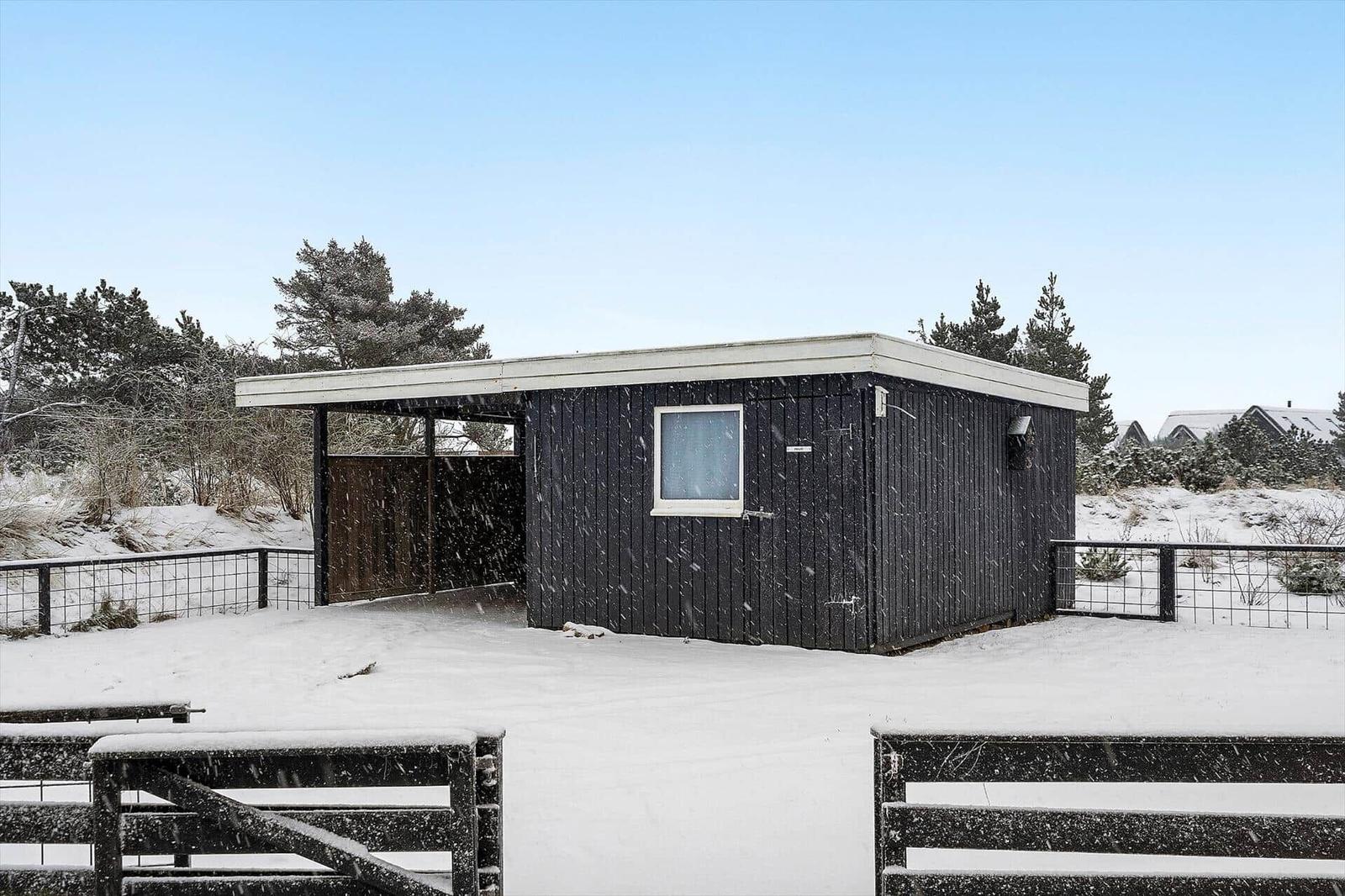 Schwarze Holzhütte mit weißem Dach und Fenster, umgeben von Schnee und Bäumen.