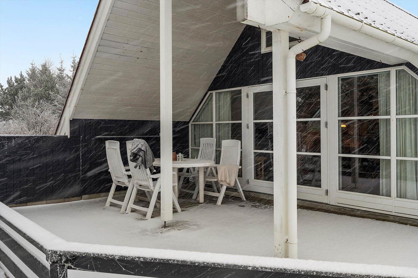 Schnee bedeckt die Terrasse mit Tisch und Stühlen vor einem Haus mit Glasfront.