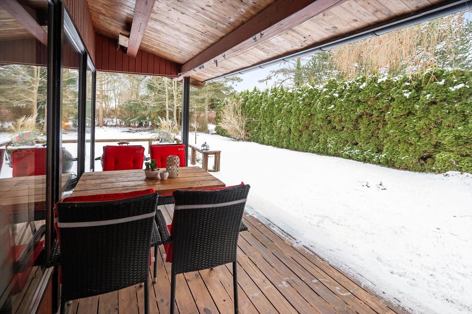 Deck with wooden table and chairs, snow in garden, green hedge in background.
