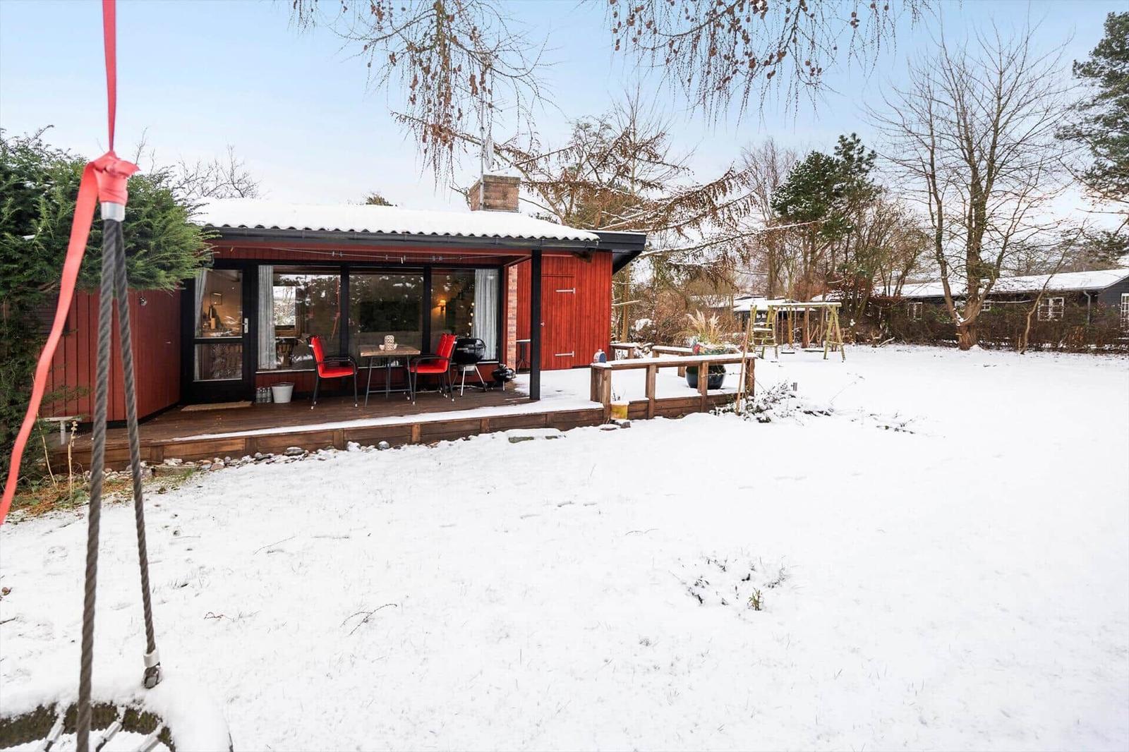 Red wooden house with terrace, snow in garden, table and chairs outside.
