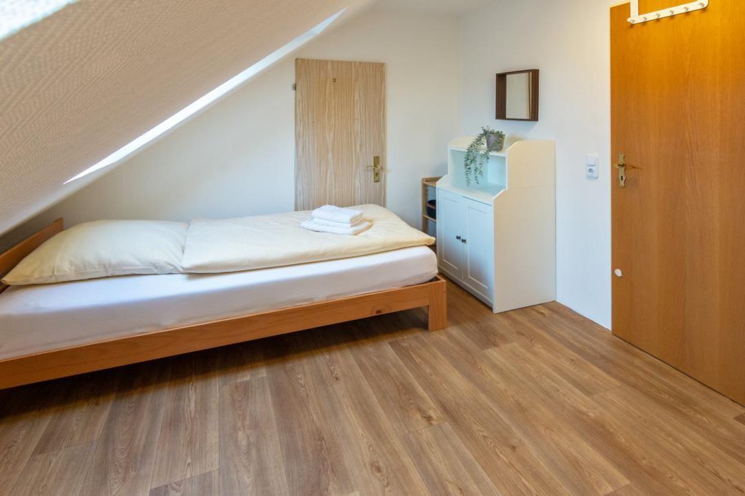 Bedroom with sloped ceiling, wooden floor, and bed. Small cabinet with plant.