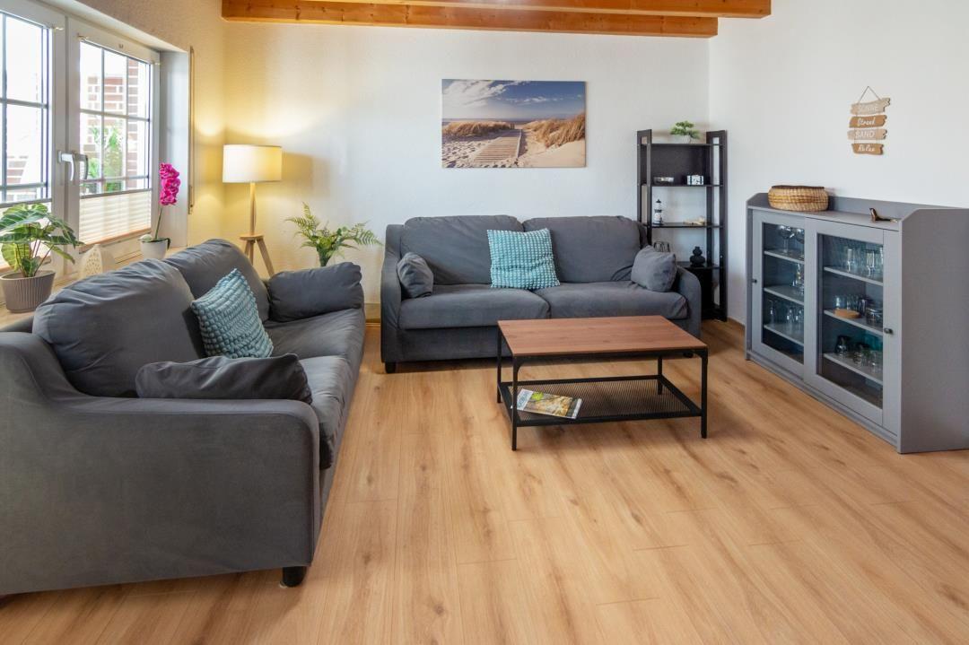 Living room with two sofas, table, and wooden floor. Wall picture and plants visible.