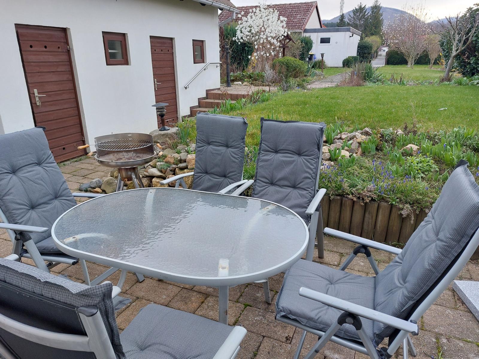 Terrasse mit Tisch, Stühlen und Grill. Hintergrund: Garten und Haus.