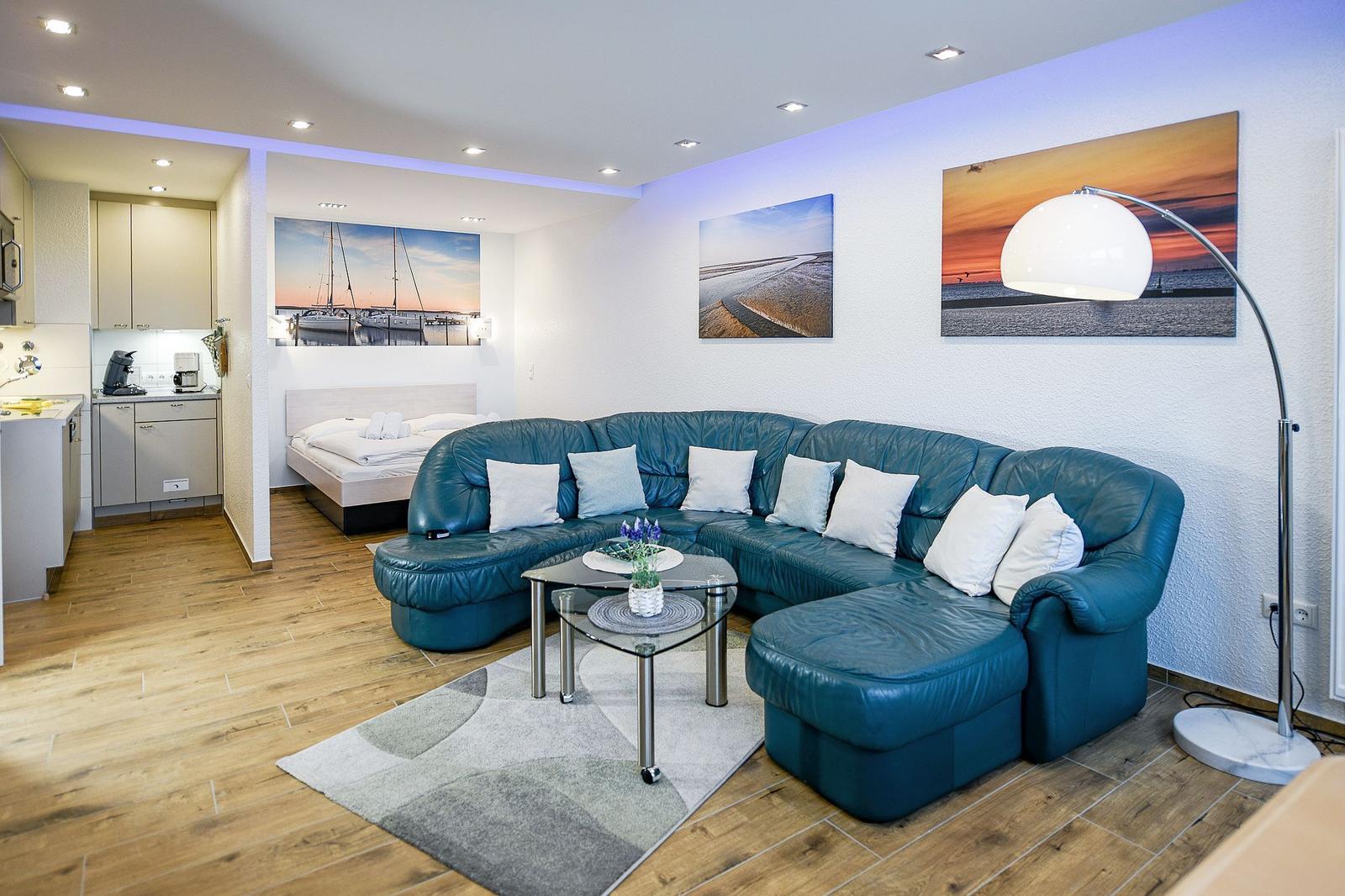 Wohnbereich mit blauem Sofa, Glas-Tisch und Küchenbereich im Hintergrund.