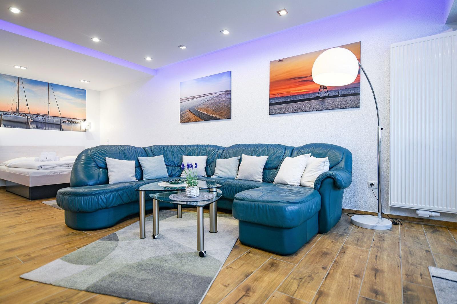 Blauer Sofa-Bereich mit Glas-Tisch und Wandbildern. Im Hintergrund ein Bett.