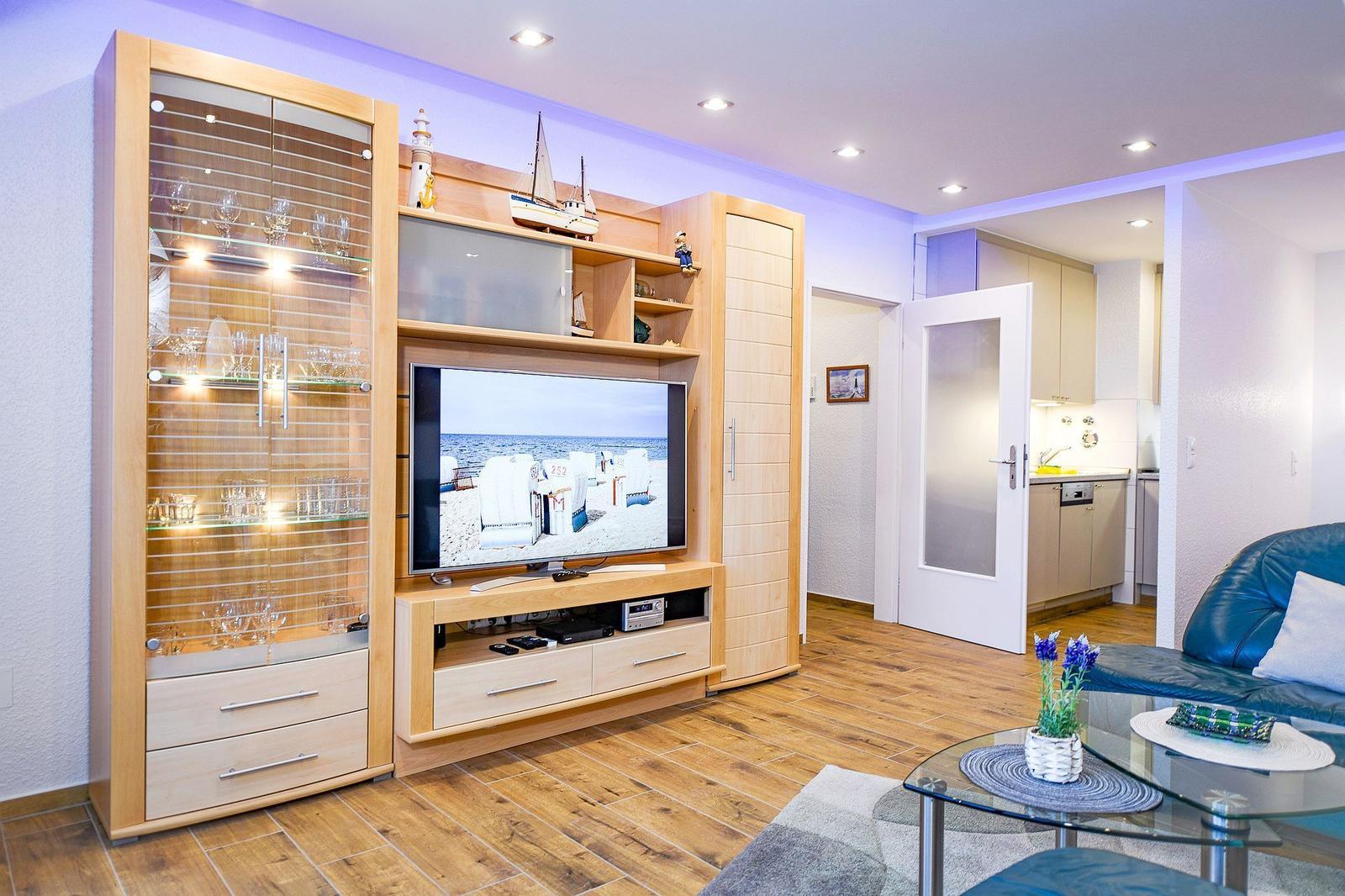 Wohnzimmer mit TV, Glasvitrine und blauem Sofa. Küche im Hintergrund sichtbar.