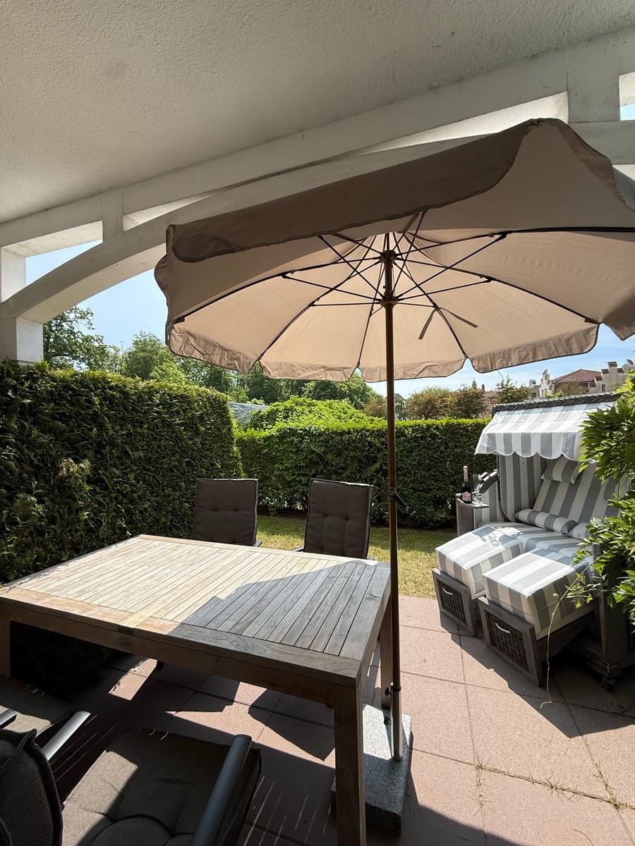 Terrasse mit Tisch, Stühlen und Sonnenschirm. Hintergrund: grüner Heckenzaun.