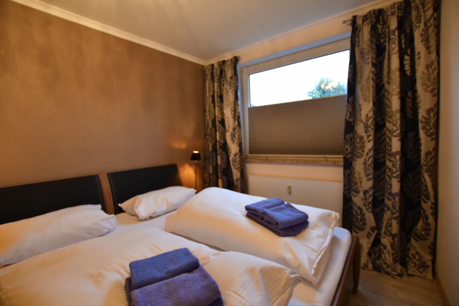 Doppelbett mit weißen Laken und blauen Handtüchern. Fenster mit floralen Vorhängen.