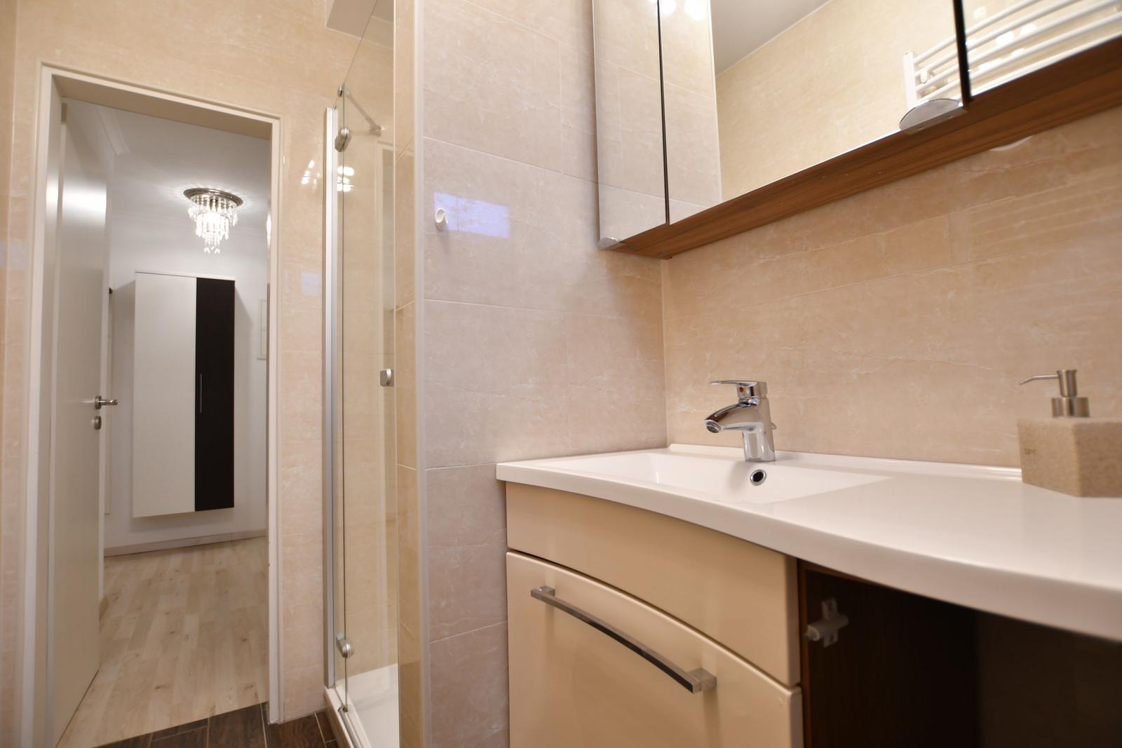 Bad mit Waschbecken, Spiegel und Dusche. Blick in angrenzenden Raum.