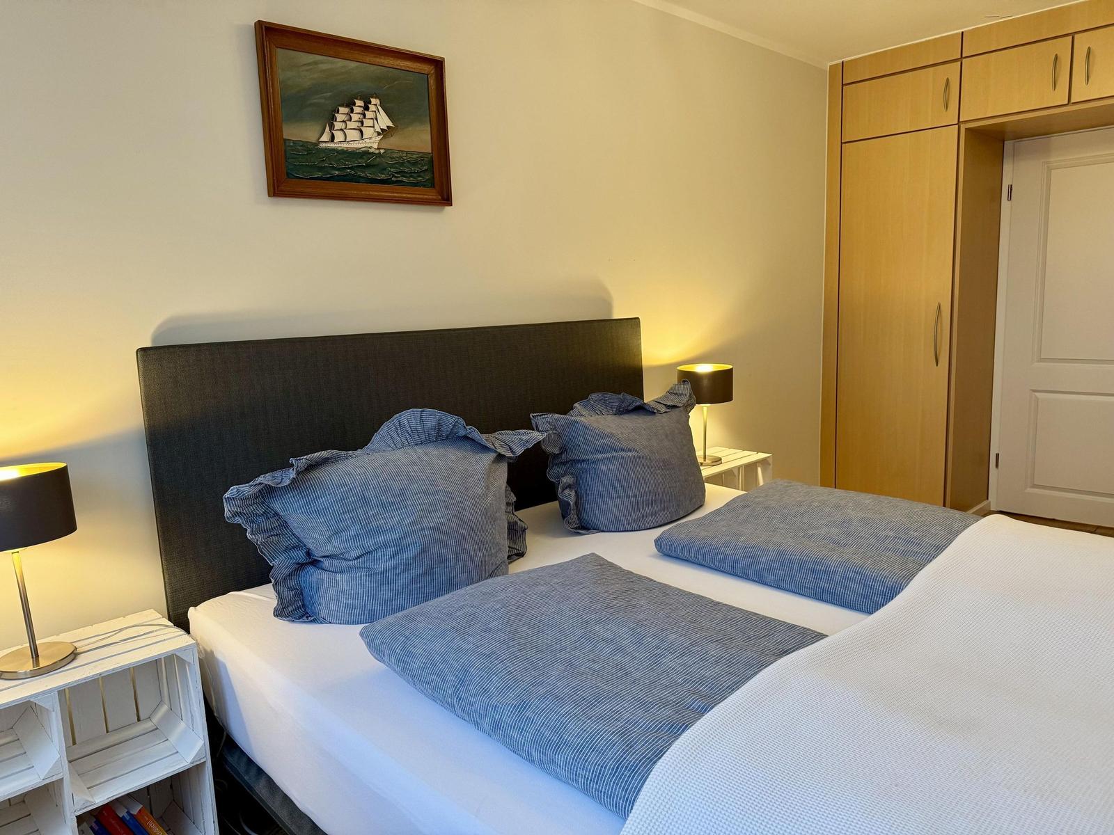 Schlafzimmer mit Bett, Nachttischen und Kleiderschrank. Wandbild zeigt Segelschiff.