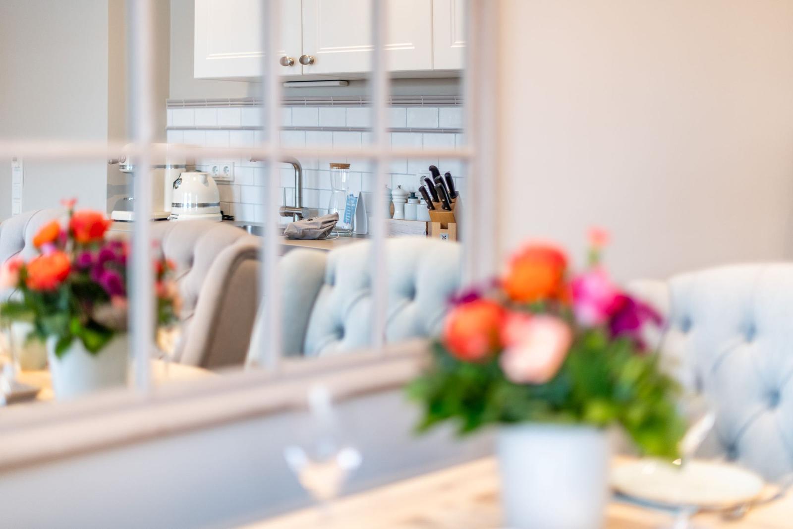 Küche mit weißen Schränken und Küchenzeile. Im Vordergrund Blumen und Stühle.