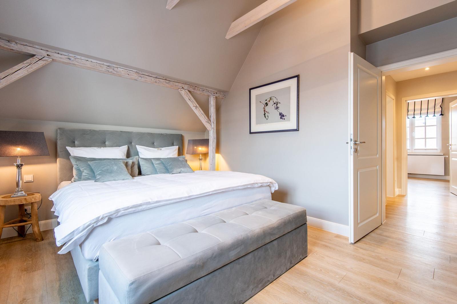 Schlafzimmer mit Doppelbett, Holzbalken und Fußbodenheizung. Tür führt in angrenzenden Raum.