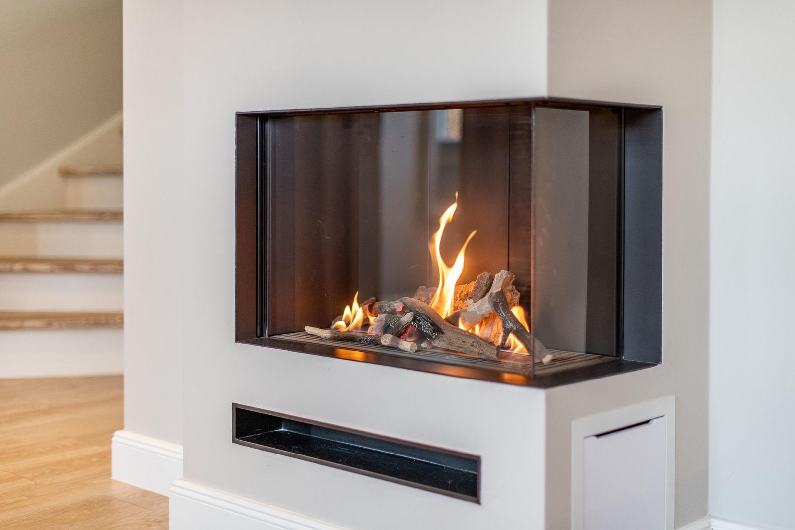 Ein moderner Kamin mit brennenden Holzscheiten in einem hellen Raum mit Holzboden und Treppe.