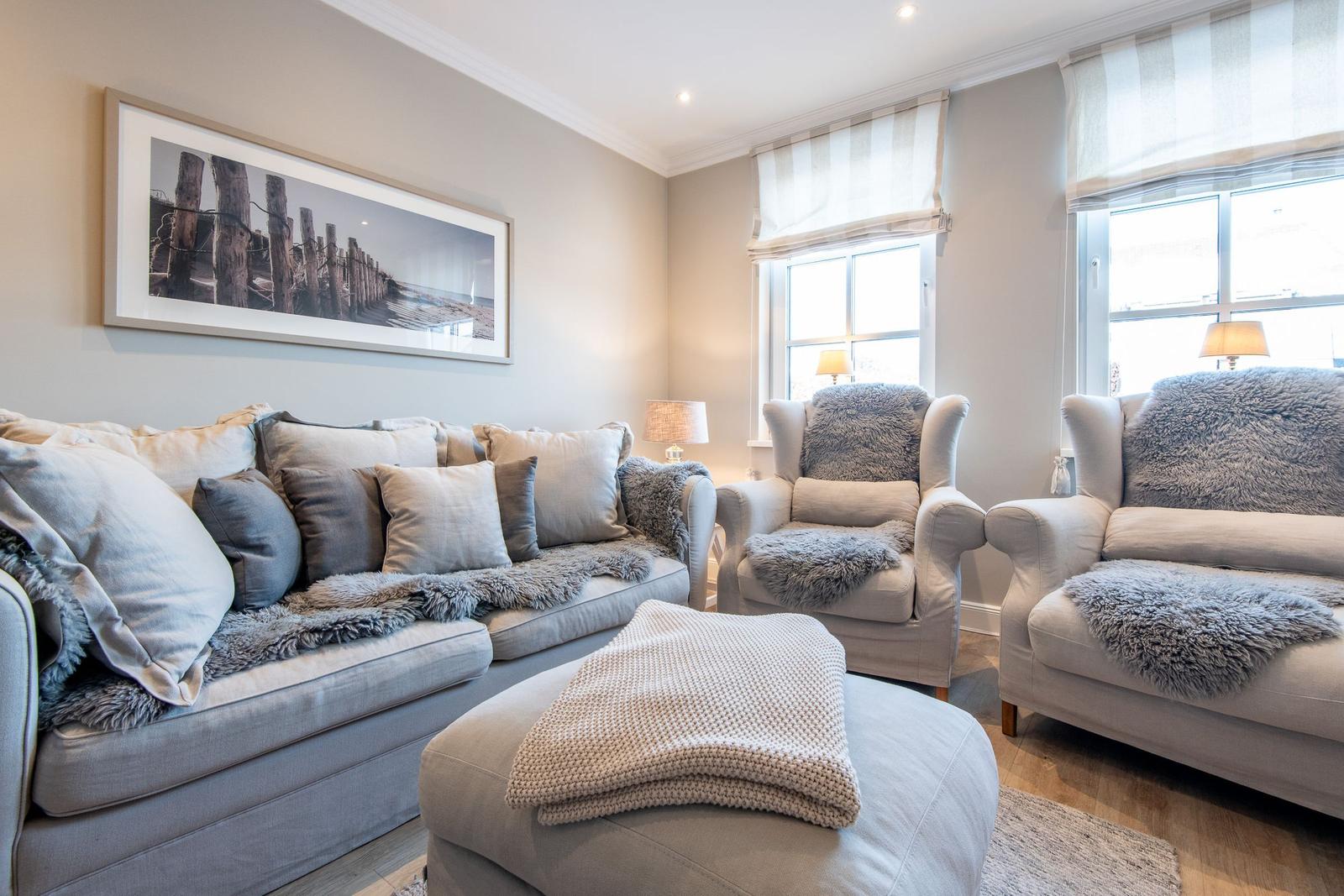 Wohnzimmer mit Sofa, zwei Sesseln und Fußhocker. Fenster mit Rollläden und Wandbild.