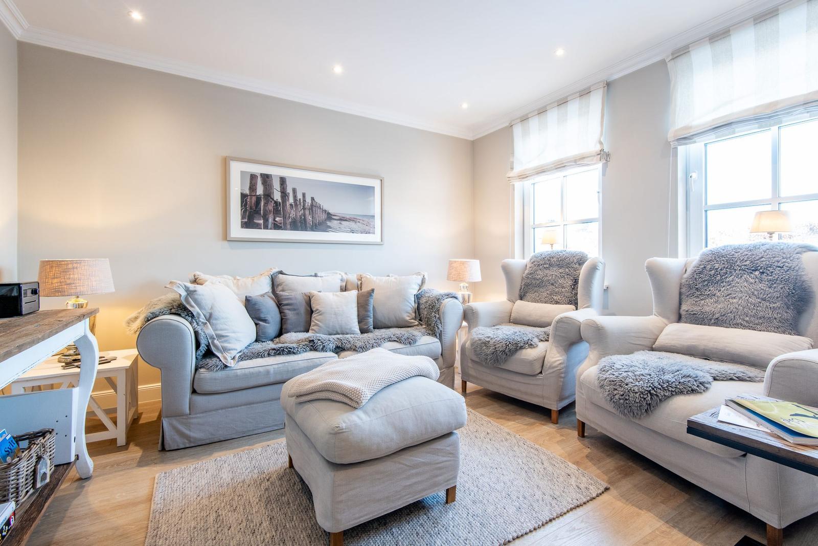 Wohnzimmer mit Sofa, zwei Sesseln, Fußhocker und Wandbild. Holzboden und Fenster mit Rollläden.