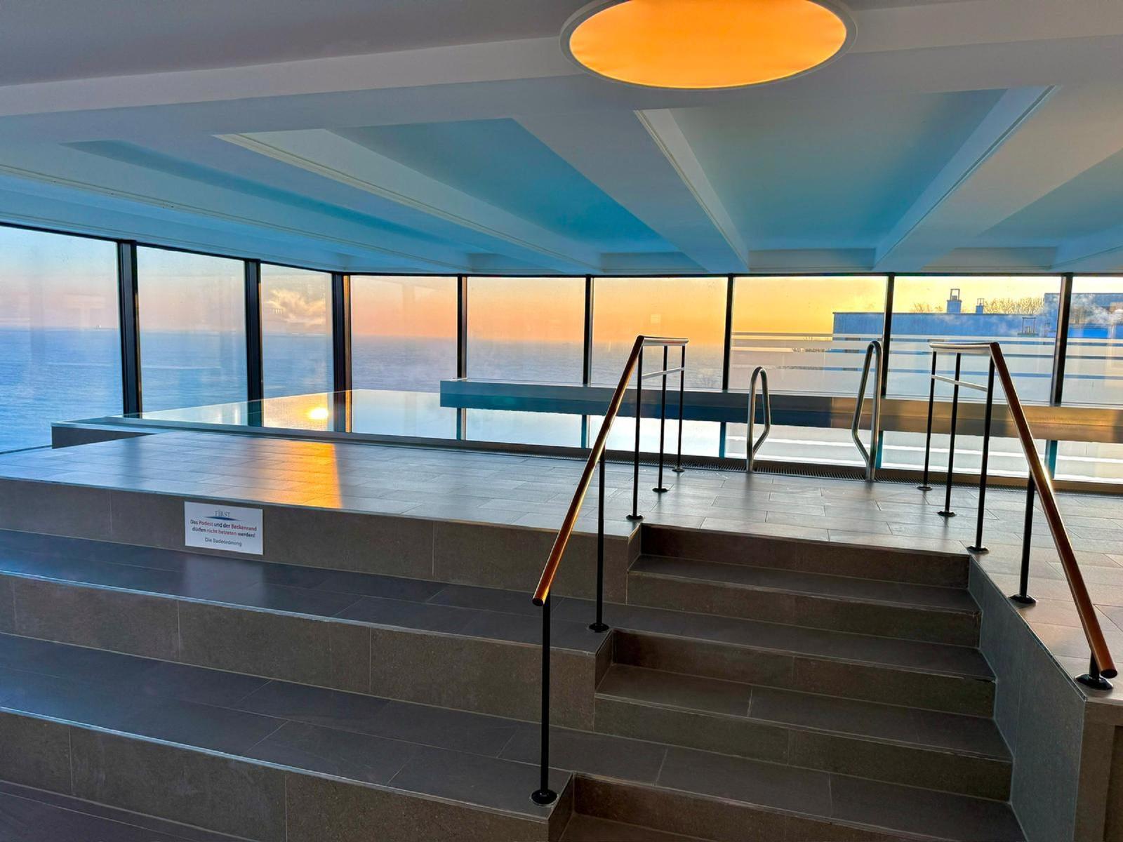 Pool mit Meerblick, Treppe und Geländer. Sonnenuntergang durch große Fenster.