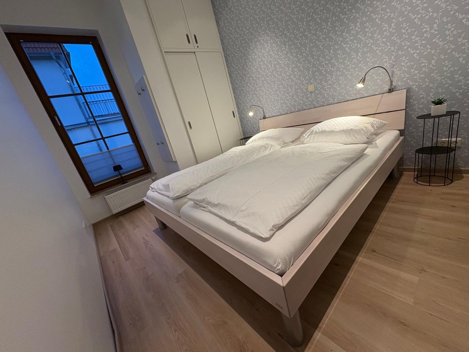 Schlafzimmer mit Doppelbett, Schrank und Fenster. Holzboden und gemusterte Tapete.