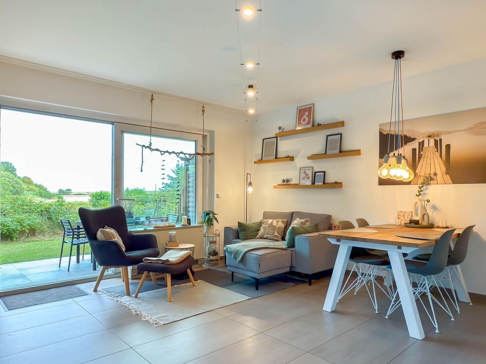 Wohn- und Essbereich mit Sofa, Stuhl und Tisch. Große Fenster mit Blick auf Garten.