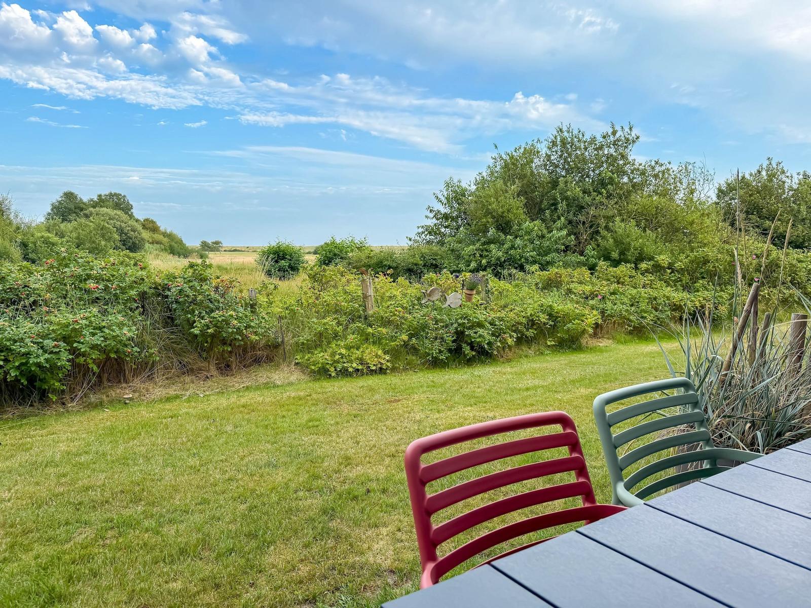 Garten mit Tisch und Stühlen, Blick auf grüne Wiesen und Bäume unter blauem Himmel.