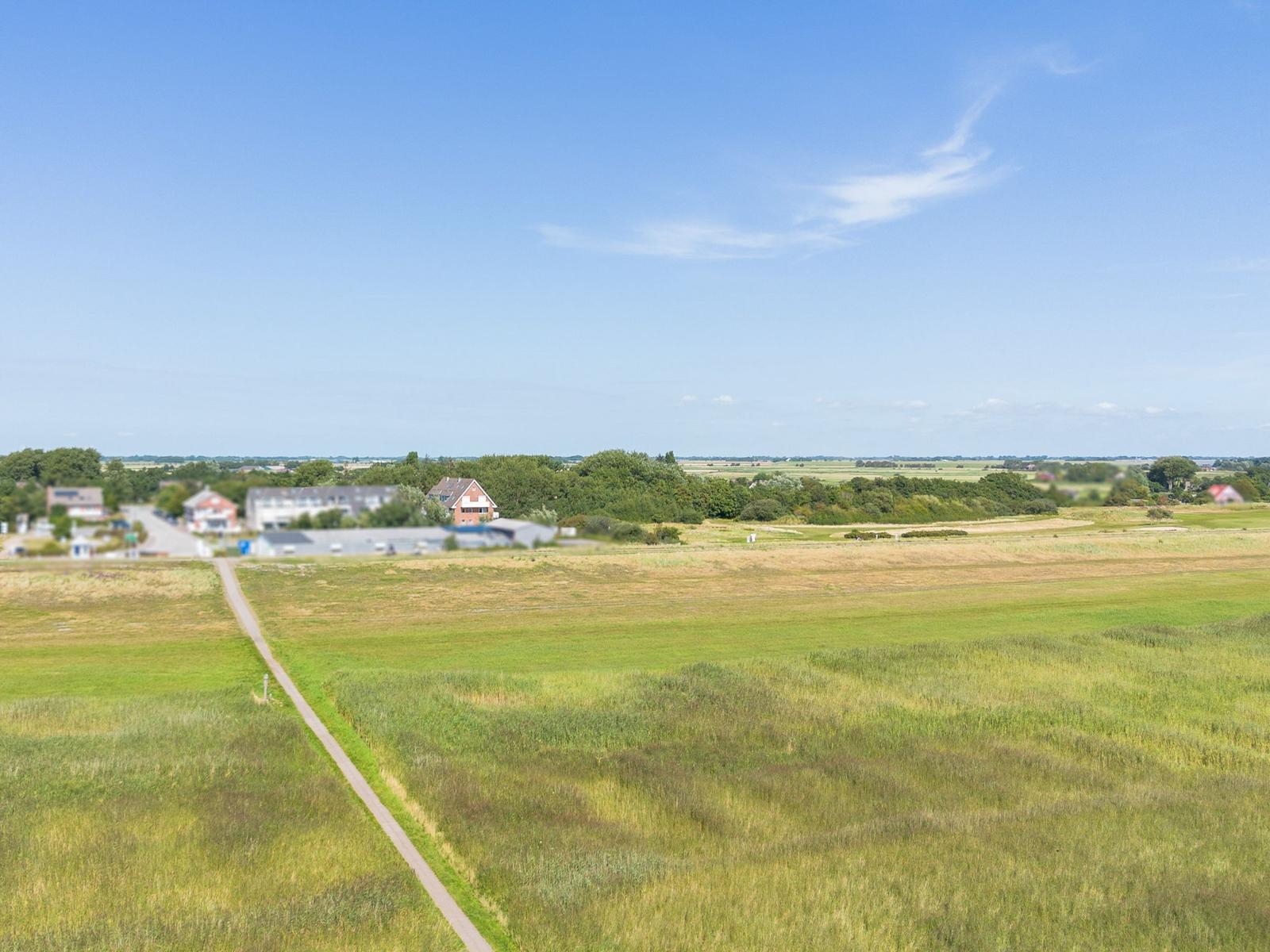 Weitläufige Grünfläche mit Weg und Ferienhaus im Hintergrund unter blauem Himmel.