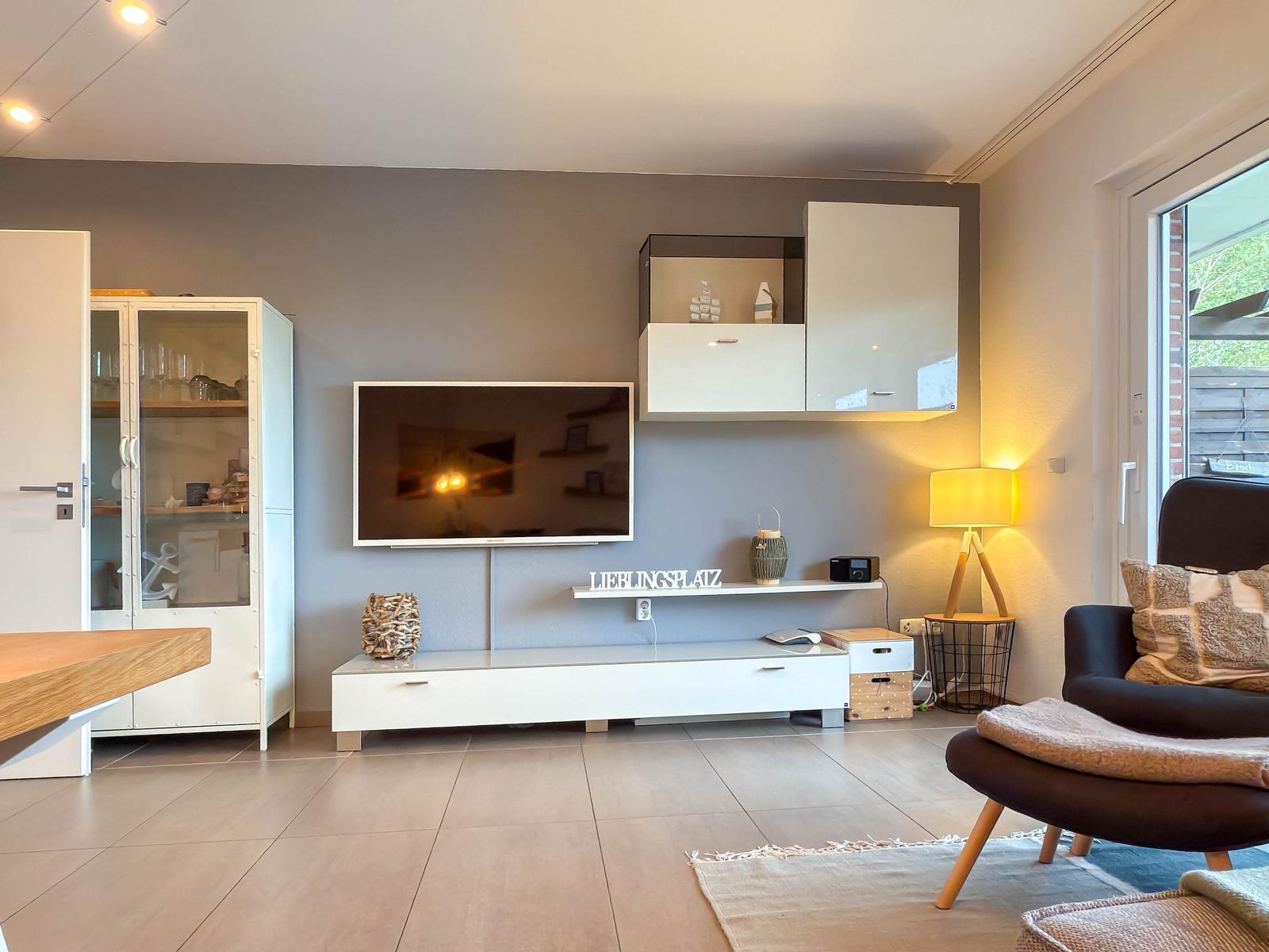 Wohnzimmer mit TV, Schränken und Sitzgelegenheit. Fenster zeigt Außenbereich.