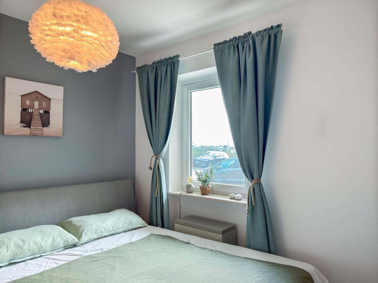 Schlafzimmer mit Bett, Fenster, Vorhängen und Deckenleuchte.