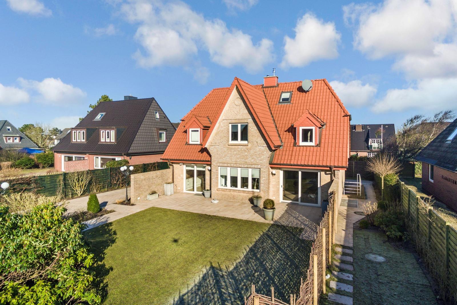 Haus mit rotem Dach, Garten und Terrasse unter blauem Himmel.
