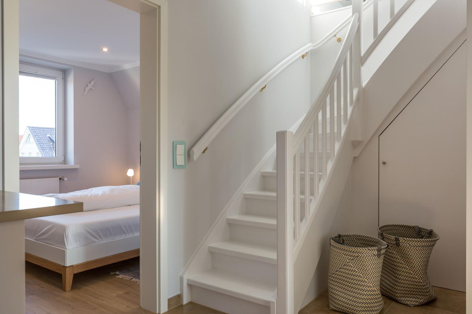 Treppenhaus mit Blick in Schlafzimmer. Weiße Treppe, zwei Körbe und Bett mit Fenster.